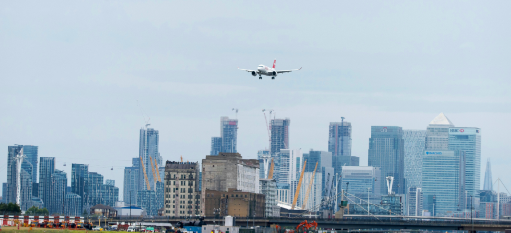 A commercial airplane descends towards a landing at an airport near London, with the city skyline and skyscrapers in the background, symbolizing the international travel and mobility impacted by the UK’s digital eVisa transition.